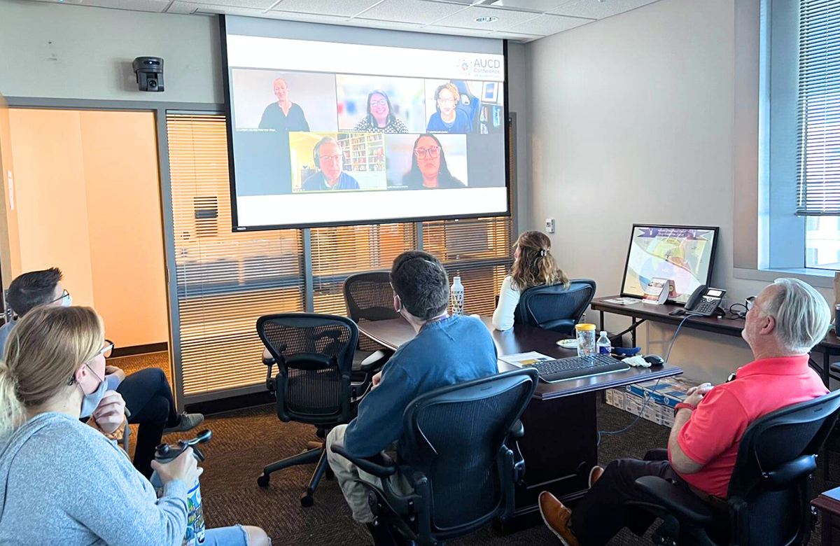 A group of people sitting in chairs in a room watching a webinar projected on  a screen.